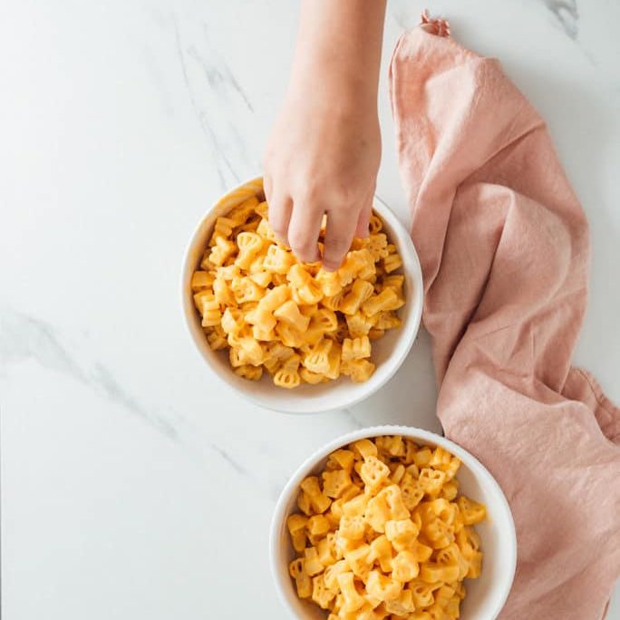 little girl hand reaching for delicious pastabilities mac and cheese on kitchen counter with pink napkin