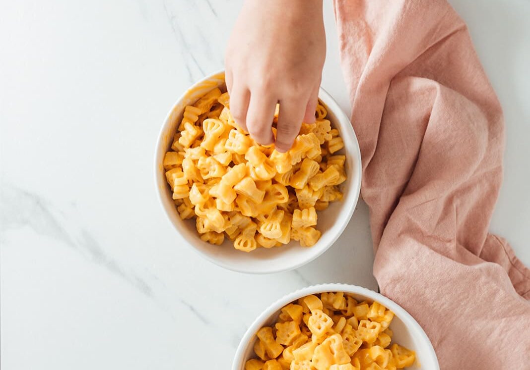 little girl hand reaching for delicious pastabilities mac and cheese on kitchen counter with pink napkin